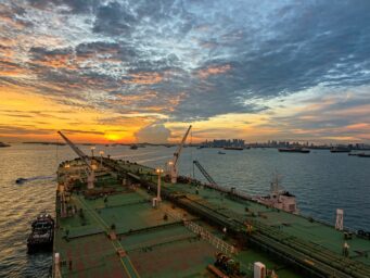 Oil tanker at sea at sunset, with other vessels and a city skyline in the background