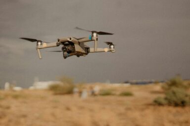A dark drone with a camera flying low over arid terrain against a grey sky