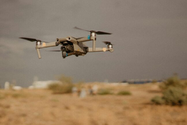 A dark drone with a camera flying low over arid terrain against a grey sky