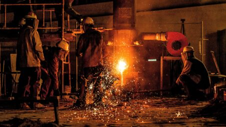 Molten steel pouring from a furnace with sparks flying, surrounded by several workers in an industrial steel plant