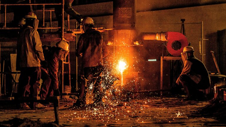 Molten steel pouring from a furnace with sparks flying, surrounded by several workers in an industrial steel plant