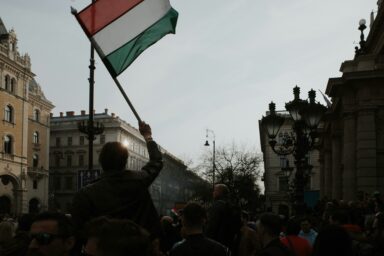 A person holds a Hungarian flag in the foreground, residential buildings visible in the background at dusk