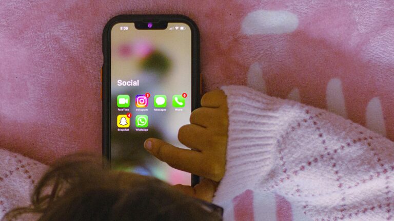 A child’s hand reaching for a social media icon on a smartphone touchscreen.