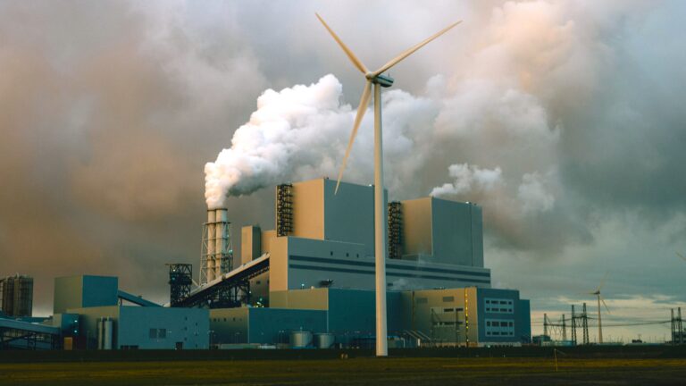 Smoke rises from the chimneys of a coal-fired power plant, with a lone wind turbine standing in front