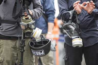 Two journalists wearing gas masks and carrying a press helmet at a public event.
