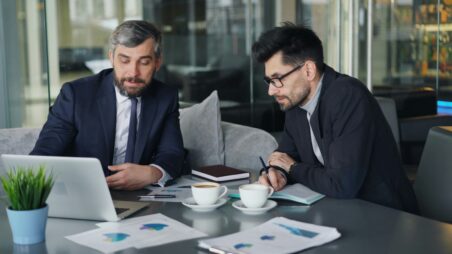 Two business executives sitting together at a conference table reviewing documents on a laptop