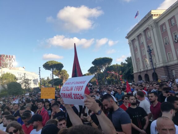 Crowd of protesters in Tirana holding Albanian flags and a sign reading "Wind of Change, Berlin 1989, Tiranë 2026"