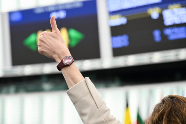 A hand giving a thumbs-up gesture during a vote in the European Parliament plenary, with a results screen visible in the background