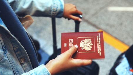 Close-up of a tourist holding a Russian passport in one hand while gripping the handle of a suitcase with the othe