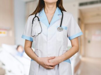 A nurse in a hospital ward with a patient in the background.