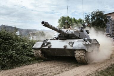A military tank in motion with a soldier visible in the hatch, dust rising behind it.