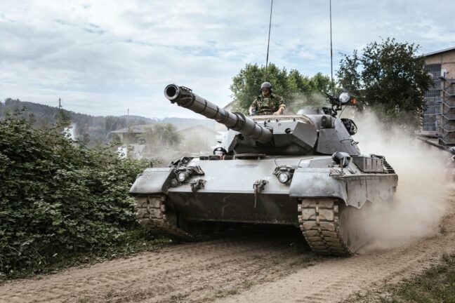 A military tank in motion with a soldier visible in the hatch, dust rising behind it.