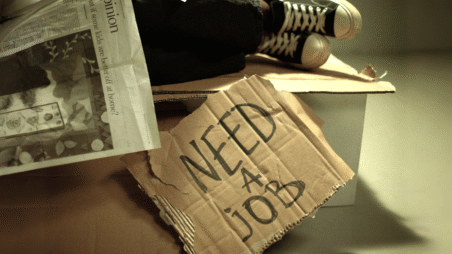 A person sleeping on a bench covered by newspapers, with a cardboard sign underneath reading “need a job”