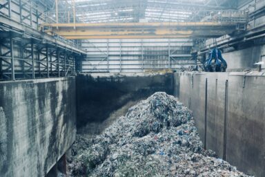 A waste processing facility with a large pile of mixed plastic waste inside an industrial hall, with a crane visible in the background.