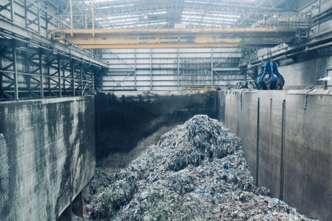 A waste processing facility with a large pile of mixed plastic waste inside an industrial hall, with a crane visible in the background.