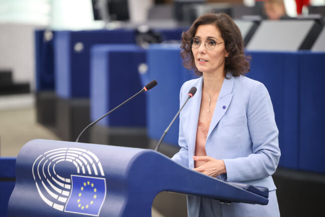Commissioner Hadja Lahbib speaks at a podium in the European Parliament chamber, with EU flag microphone stand visible.
