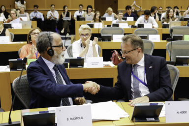 Two men shaking hands at a committee meeting table, with other MEPs seated in the background