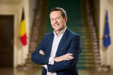 Michel De Maegd standing with arms crossed in front of a staircase, with the Belgian and European Union flags behind him