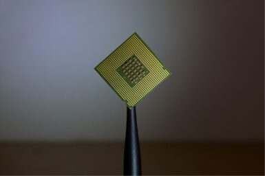 A computer processor chip with gold pins, held up against a blurred grey background.