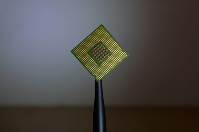 A computer processor chip with gold pins, held up against a blurred grey background.