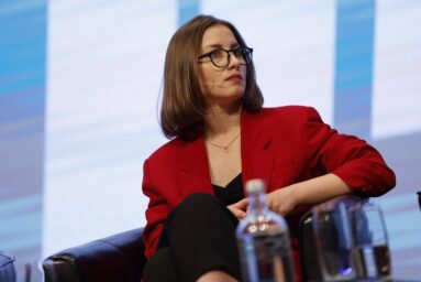 Anastasia Burakova, wearing glasses and a red blazer, sits on stage during a panel discussion.
