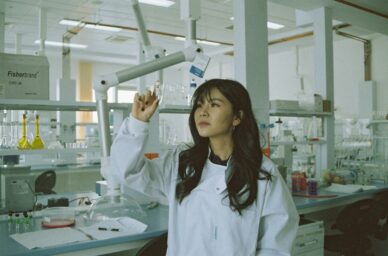 A young woman in a white lab coat holds up a small glass vial in a research laboratory, examining it closely against the light.