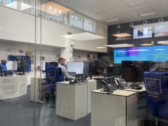 Staff at desks inside the EU's Emergency Response Coordination Centre, with large monitoring screens showing disaster maps in the background