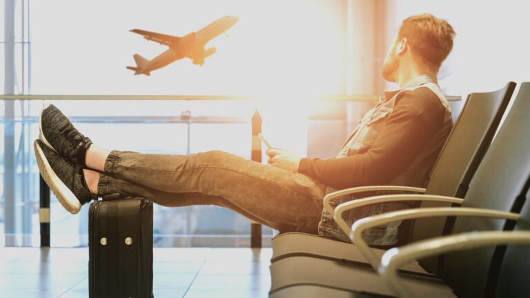 Traveller sitting at an airport with his feet on his suitcase, watching a plane take off