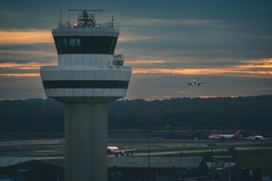 Air traffic control tower at dusk with an aircraft approaching to land and two more aircraft on the runway below.