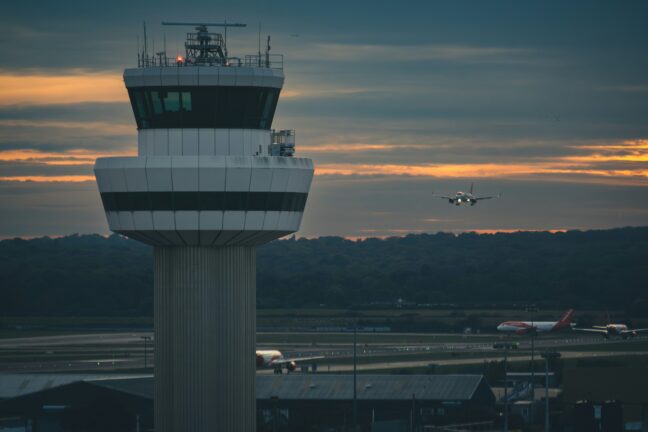 Air traffic control tower at dusk with an aircraft approaching to land and two more aircraft on the runway below.