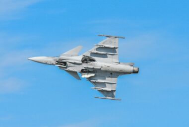 A Swedish Air Force Gripen fighter jet banking against a clear blue sky, its underwing weapons pylons visible.