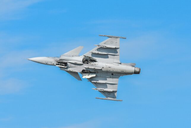 A Swedish Air Force Gripen fighter jet banking against a clear blue sky, its underwing weapons pylons visible.