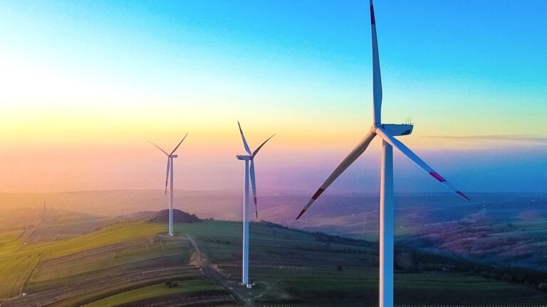 Three wind turbines on a hill silhouetted against a rainbow-colored sky at sunset