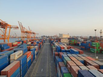 Shipping containers stacked at a port, with cranes in the background at dusk