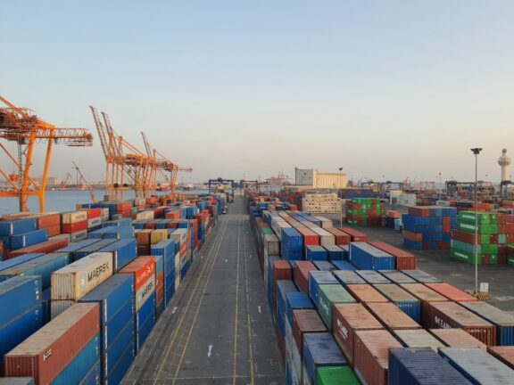 Shipping containers stacked at a port, with cranes in the background at dusk