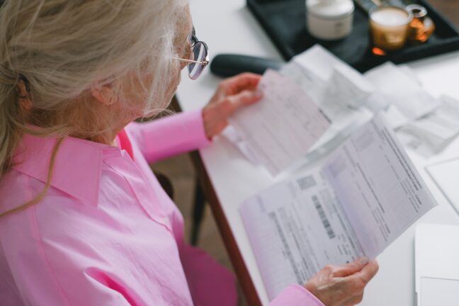 An older woman in a pink shirt reviewing paper energy bills at a table
