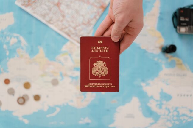 A hand holds a red Russian Federation passport upside down over a world map, with coins and a camera nearby.