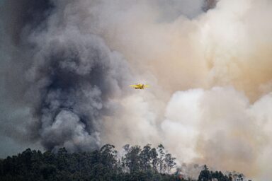 A yellow firefighting aircraft flies past a massive column of smoke rising from a forest fire