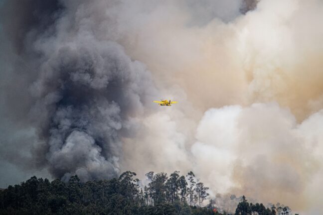 A yellow firefighting aircraft flies past a massive column of smoke rising from a forest fire