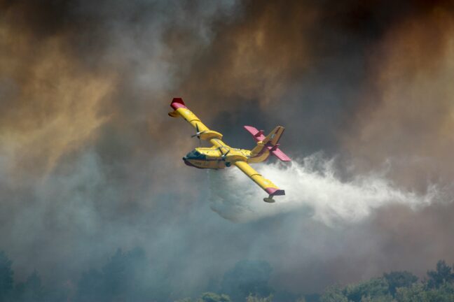 A yellow and red firefighting aircraft dropping water over a smoke-covered forest