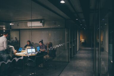 Colleagues in a glass-walled meeting room look together at a laptop screen