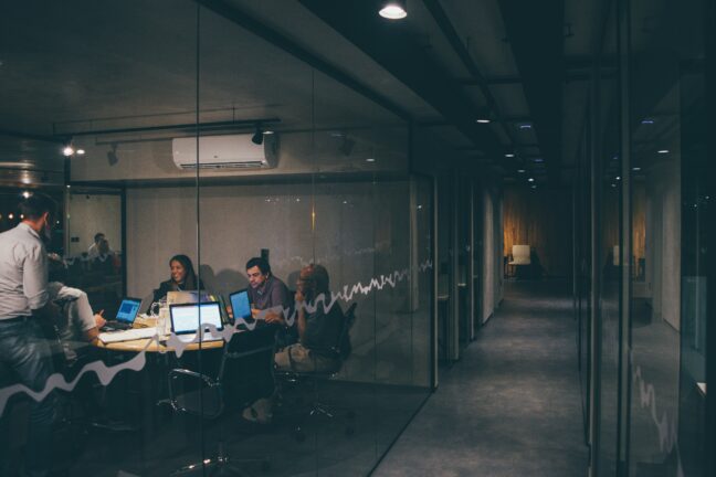 Colleagues in a glass-walled meeting room look together at a laptop screen