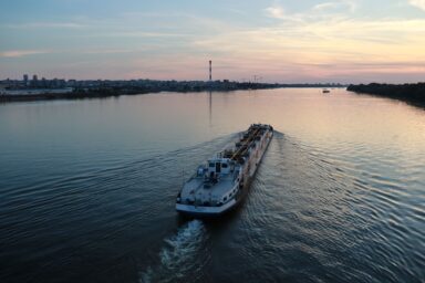 Cargo ship Srbija Beograd sailing on the Danube past Belgrade at dusk, city skyline in the background.