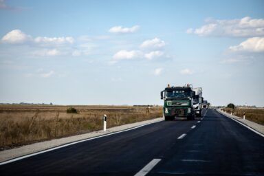 A green Volvo lorry and other trucks driving on an open road past fields, under a blue sky with clouds