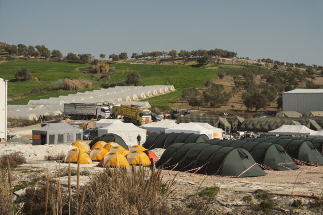 Rows of tents in a temporary camp, set up on open ground near hills and greenhouses.