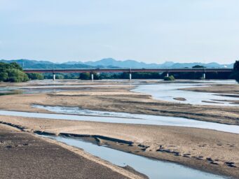 A bridge crossing a wide river with an exposed, cracked riverbed and mountains in the background
