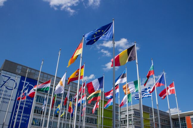 Flags of European Union member states on flagpoles against a blue sky, with the EU flag flying above them
