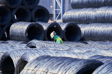 A worker in a hard hat and high-visibility vest stands among towering stacks of coiled steel wire in an industrial yard.
