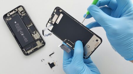 Two gloved hands use a screwdriver to repair the internal components of a smartphone, with its battery and spare parts laid out beside it.