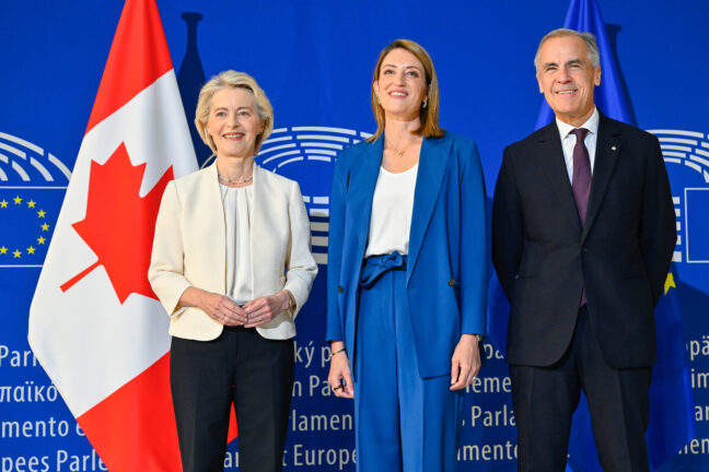 Ursula von der Leyen, Roberta Metsola and Mark Carney standing together in front of Canadian and EU flags at the European Parliament